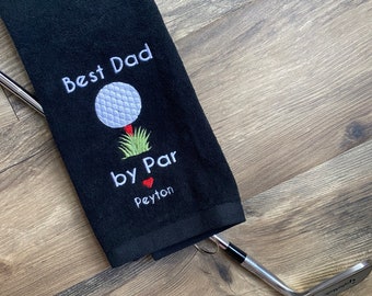 May include: Black golf towel with white embroidery that reads "Best Dad by Par" with a red heart and the name Peyton. A golf club is lying on top of the towel.