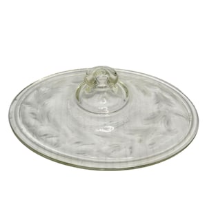 May include: An oval clear glass lid featuring a central knob handle and a decorative white frosted swirl pattern across the surface. The lid has a smooth, rounded edge and a transparent finish, designed to fit a corresponding oval container.