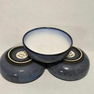 May include: Three blue ceramic bowls with a gradient effect, transitioning from white to blue. The bowls have a dark blue rim and a black base. The bowls are stacked together, with the bottom of two bowls visible.