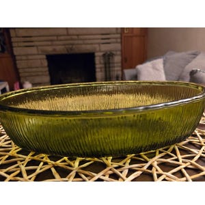 May include: An oval-shaped, olive-green glass bowl with a textured, vertical-line pattern. The bowl sits on a woven, light-brown placemat. The background includes a fireplace and a glimpse of a living room setting.