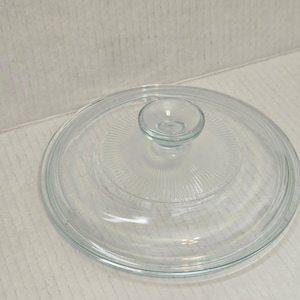 Op de afbeelding: Een doorzichtige glazen deksel met een kleine, afgeronde handgreep. De deksel heeft een cirkelvormig ontwerp met concentrische ribbels die vanuit het midden uitstralen. Het glas is helder, waardoor je de inhoud kunt zien.