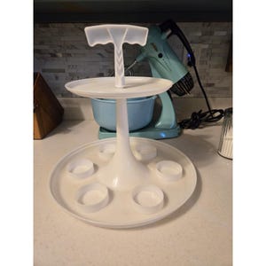 Könnte beinhalten: Ein weißes Serviertablett aus Kunststoff mit zwei Ebenen und sechs kleinen Becherhaltern auf der unteren Ebene. Die obere Ebene hat einen Griff. Das Tablett ist zum Servieren von Vorspeisen oder Desserts konzipiert.