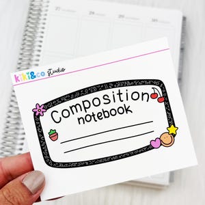 May include: A white composition notebook sticker with the text "Composition notebook" in black. The sticker has a black border with decorative elements like a flower, cherry, and star. The sticker is held in a hand.