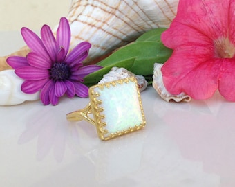 May include: A gold ring with a square white opal gemstone set in a gold filigree frame. The ring is on a white surface with pink and purple flowers and seashells.
