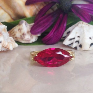 Op de afbeelding: Een gouden ring met een grote, helderrode marquise-vormige edelsteen. De ring is geplaatst tegen een achtergrond van schelpen en paarse bloemen.