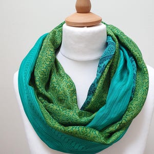 May include: An infinity scarf featuring a layered design with a vibrant teal solid fabric and a contrasting green fabric adorned with an intricate, repeating paisley-inspired floral pattern in shades of lime and forest green throughout the textile.