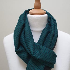Peut inclure: Écharpe à motifs teal et noirs drapée sur un mannequin blanc. L'écharpe présente un motif répété de petites formes noires sur un fond teal. L'écharpe est faite d'une matière douce et est présentée sur un mannequin blanc.