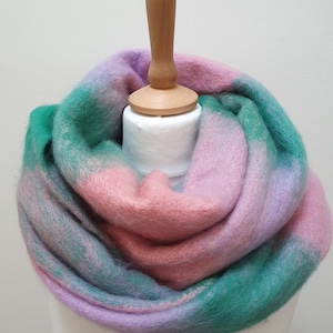 May include: A soft, fuzzy infinity scarf featuring a blended, variegated pattern of pastel pink, lavender, and deep teal green. The scarf is draped in multiple loose, voluminous folds, showcasing a thick, brushed texture throughout the fabric.