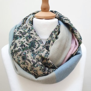 Peut inclure: Une écharpe douce et confortable avec un motif paisley dans des tons de bleu, vert et rose. L'écharpe est enroulée autour du cou d'un mannequin et a un fond gris clair.