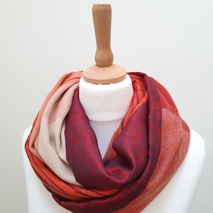 Op de afbeelding: Een veelkleurige infinity sjaal, gepresenteerd op een witte mannequin. De sjaal heeft tinten rood, oranje en crème. De sjaal heeft een bloemenpatroon in het bordeauxrode gedeelte. De sjaal is gemaakt van een zacht materiaal.