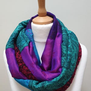 May include: An infinity scarf featuring a patchwork design of purple satin, teal fabric with a black paisley pattern, and a textured dark red knit material. The scarf is draped in a circular loop, showcasing the contrasting colours and textures.