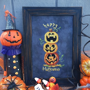 May include: A framed cross-stitch design featuring a stack of three jack-o'-lantern pumpkins with the words "happy halloween" on a dark blue background.