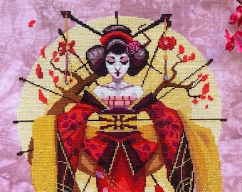May include: Cross-stitch artwork of a geisha in a red kimono with floral accents, set against a pink background. The geisha is framed by a gold border and adorned with branches of pink blossoms. A yellow sash flows from the kimono.