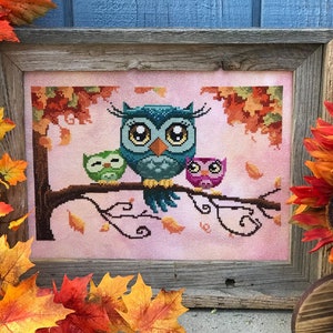 Puede incluir: Una bordado de punto de cruz con tres búhos posados en una rama con hojas de otoño. Los búhos son azules, verdes y rosas con ojos grandes. El fondo es de color rosa suave con hojas que caen.