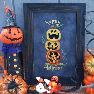 May include: A framed cross-stitch design featuring a stack of three jack-o'-lantern pumpkins with the words "happy halloween" on a dark blue background.