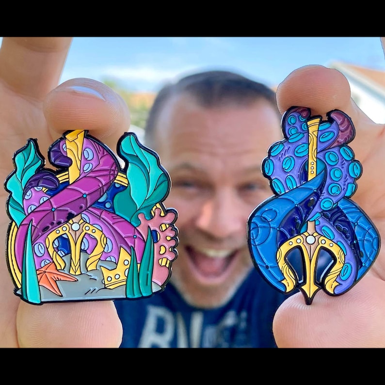 May include: Two enamel pins featuring an octopus and a trident. The octopus pin is purple, blue, and gold. The trident pin is blue, gold, and pink. Both pins have a detailed design.