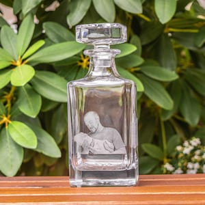 May include: A clear glass decanter with a square body and a stopper. The decanter has a photo of a person holding a small child etched into the glass.