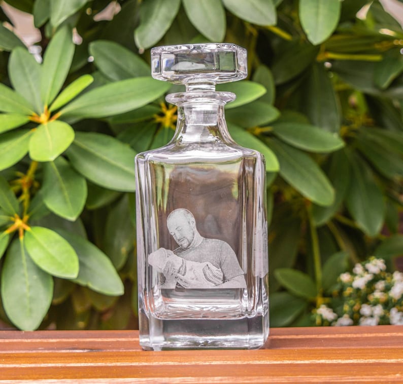 May include: A clear glass decanter with a square body and a stopper. The decanter has a photo of a person holding a small child etched into the glass.