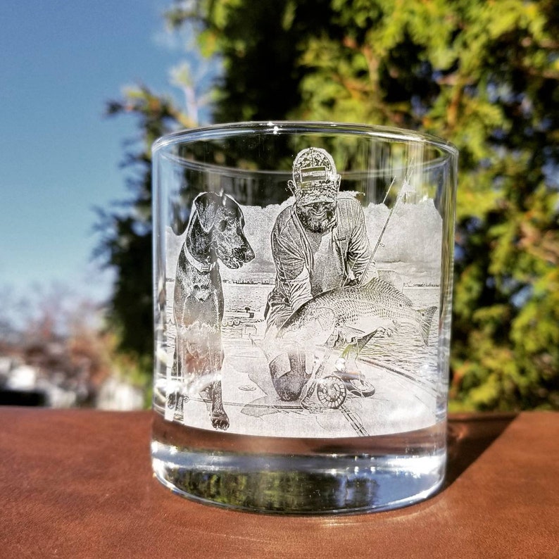 Puede incluir: Un vaso de vidrio transparente con una imagen grabada de una persona sosteniendo un pez y un perro de pie a su lado. La imagen est&aacute; en blanco y negro.