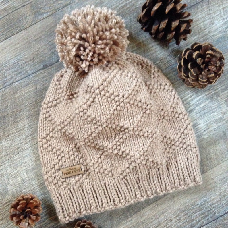 Op de afbeelding: Een beige gebreide beanie met een pompon bovenop en een bruin leren patch met de tekst "The Vintage Pine Cone".