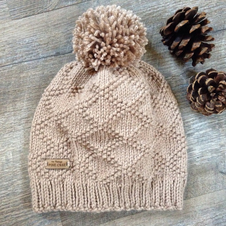 Op de afbeelding: Een beige gebreide beanie met een pompon bovenop. De beanie heeft een ruitpatroon en een bruin lederen label met de tekst "The Vintage Pine Cone".