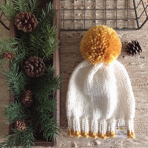 May include: White knitted beanie with a yellow pom pom and a yellow trim. The beanie has a label that reads "The Vintage Pure Cone".
