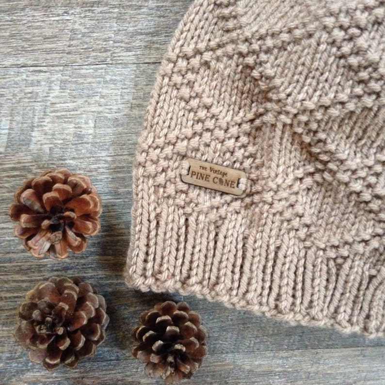 Op de afbeelding: Een beige gebreide beanie met een bruin leren label waarop "The Vintage Pine Cone" staat.