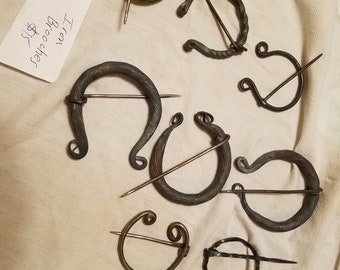 May include: A collection of eight iron brooches, some with intricate designs, displayed on a white fabric background. The text "Iron Brooches $12" is visible in the image.