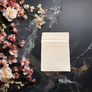 May include: A white cotton drawstring pouch with a white fabric lining. The pouch is lying flat on a dark gray and black marble surface.