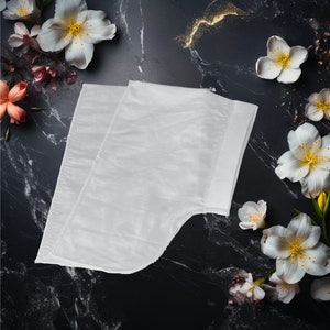 May include: White cotton fabric folded in half, with a smooth, soft texture. The fabric is laid out on a black marble surface with white flowers scattered around it.