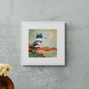May include: A framed print of a colorful painting of a kingfisher perched on a branch. The painting is set against a light green background. The frame is white and has a simple design.