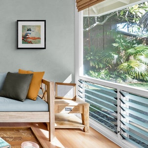 May include: A light blue sofa with a gray and orange pillow, a wooden side table with a framed picture, and a window with a view of lush green tropical plants.