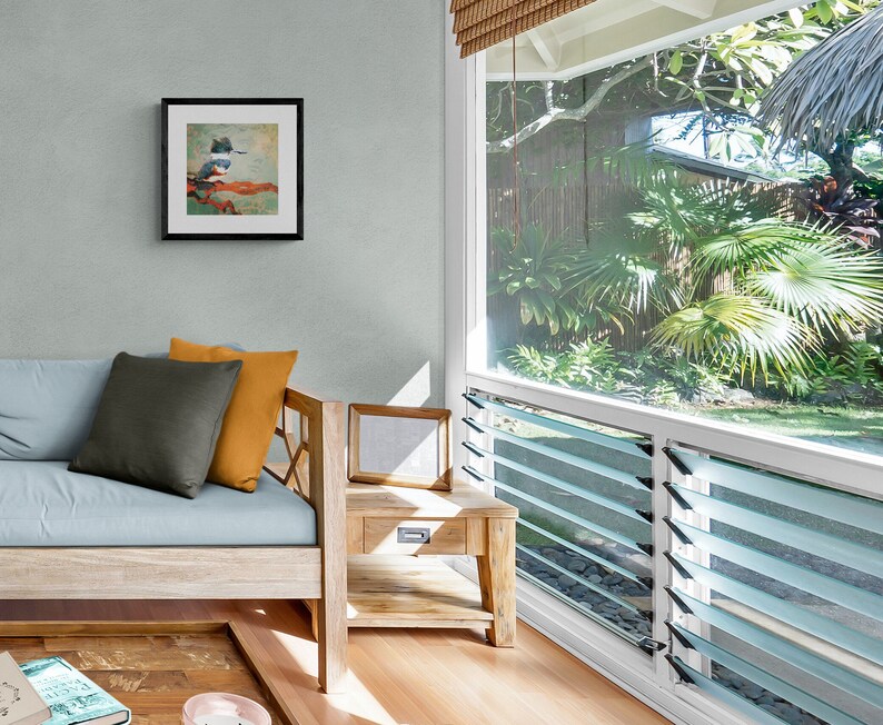 May include: A light blue sofa with a gray and orange pillow, a wooden side table with a framed picture, and a window with a view of lush green tropical plants.