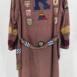 May include: A burgundy wool robe with a large blue "R" on the back. The robe is decorated with various patches, including a star, a circle with the word "Riverside" and a star, and several crests. The robe has a belt with a striped pattern and a buckle.