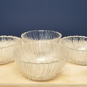 May include: Four clear glass bowls with a textured, ridged design. The bowls have a spiral pattern and a scalloped rim. They are arranged on a light-colored wooden surface against a dark blue background. These bowls are suitable for serving salads or desserts.