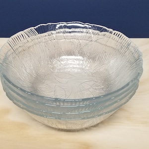May include: Three clear glass bowls with a textured leaf design, stacked on a light wood surface. Each bowl has a scalloped edge. The bowls are made of clear glass.