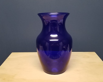 May include: A tall, cobalt blue glass vase with a flared top. The vase is cylindrical with a rounded base and sits on a light-colored wooden surface. The background is a solid, dark blue.