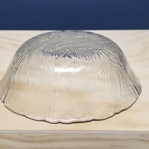 May include: A clear glass bowl with a textured, leaf-like pattern. The bowl is upside down, resting on a light-coloured wooden surface. The background is a solid dark blue.