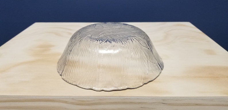 May include: A clear glass bowl with a textured, leaf-like pattern. The bowl is upside down, resting on a light-coloured wooden surface. The background is a solid dark blue.