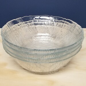 May include: Four clear glass bowls with a textured pattern, stacked on a light wooden surface. The bowls have a leaf-like design. The background is a dark blue.