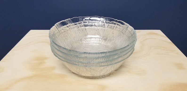 May include: Four clear glass bowls with a textured pattern, stacked on a light wooden surface. The bowls have a leaf-like design. The background is a dark blue.