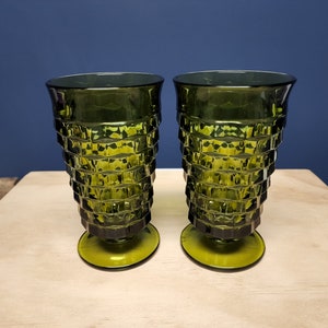 May include: Two matching olive green glass tumblers featuring a faceted, geometric diamond-cut pattern on the exterior. Each glass has a wide, flared rim that tapers down to a circular pedestal base, creating a distinct, textured silhouette.