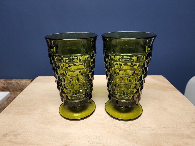 May include: Two matching olive green glass tumblers featuring a faceted, geometric diamond-cut pattern on the exterior. Each glass has a wide, flared rim that tapers down to a circular pedestal base, creating a distinct, textured silhouette.