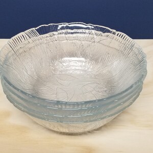 May include: Four clear glass bowls with a leaf pattern, stacked on a light wood surface. The bowls have a scalloped edge. The background is a dark blue. Each bowl is approximately 20 cm in diameter.