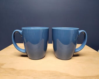 May include: Two matching blue ceramic mugs with curved handles. The mugs have a glossy finish and sit on a light brown wooden surface. The background is a dark blue wall.