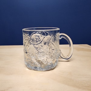 May include: A clear glass mug featuring a raised, embossed design of a character wearing a bowler hat and a polka-dot patterned suit. The mug has a smooth, curved handle on the right side and a cylindrical body with a textured, relief-style surface.