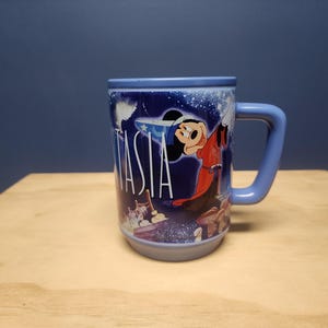 Puede incluir: Una taza de cer&aacute;mica azul con Mickey Mouse de Fantas&iacute;a. La taza tiene un asa curva y la palabra "Fantas&iacute;a" en blanco. El dise&ntilde;o incluye im&aacute;genes de Mickey Mouse con una t&uacute;nica roja y un fondo estrellado.