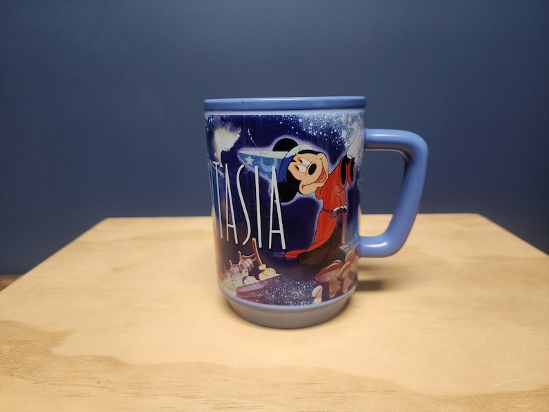 Puede incluir: Una taza de cer&aacute;mica azul con Mickey Mouse de Fantas&iacute;a. La taza tiene un asa curva y la palabra "Fantas&iacute;a" en blanco. El dise&ntilde;o incluye im&aacute;genes de Mickey Mouse con una t&uacute;nica roja y un fondo estrellado.