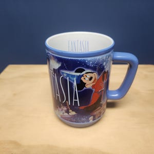 Puede incluir: Taza de cer&aacute;mica azul con Mickey Mouse de Fantasia. La taza tiene un asa y borde azules, con la palabra "Fantasia" en blanco. El dise&ntilde;o incluye im&aacute;genes de Mickey Mouse y una escena de noche estrellada.