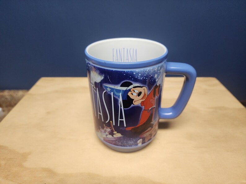 Puede incluir: Taza de cer&aacute;mica azul con Mickey Mouse de Fantasia. La taza tiene un asa y borde azules, con la palabra "Fantasia" en blanco. El dise&ntilde;o incluye im&aacute;genes de Mickey Mouse y una escena de noche estrellada.